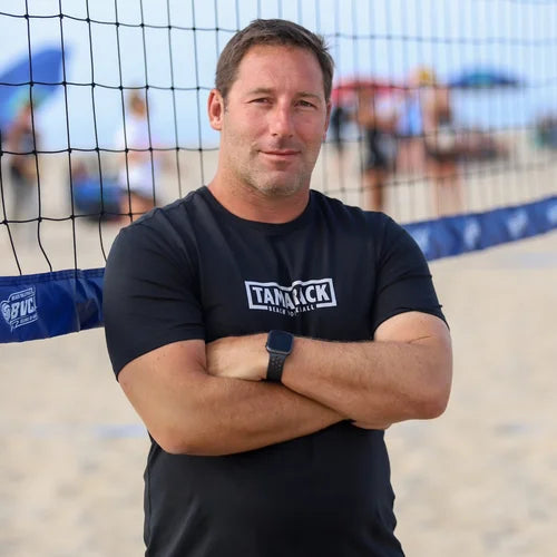 Man wearing a black 'Tamaack' t-shirt standing on a beach volleyball court.